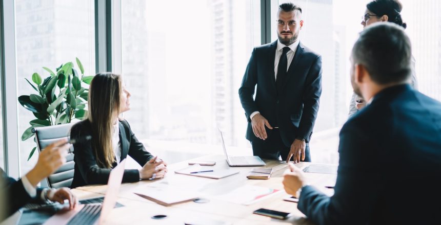 Concentrated executive male in formal clothing having discussion with board of directors while standing near window of modern conference room